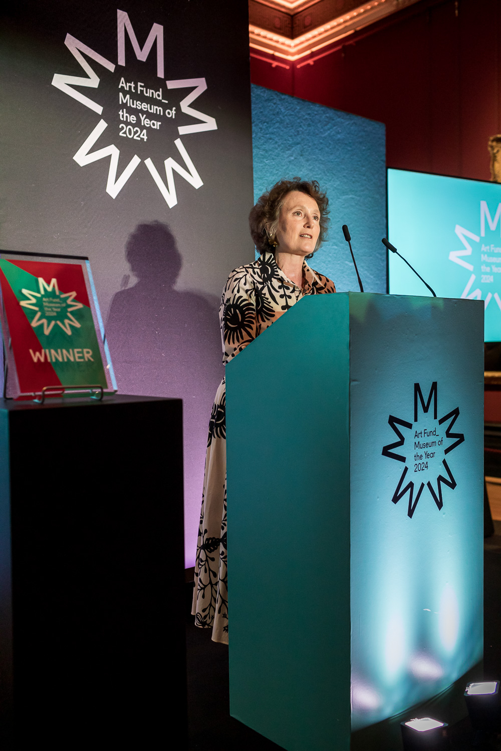 A speaker at the lectern at Museum of the Year 2024, with the starburst identity projected behind her and the winner's trophy alongside.