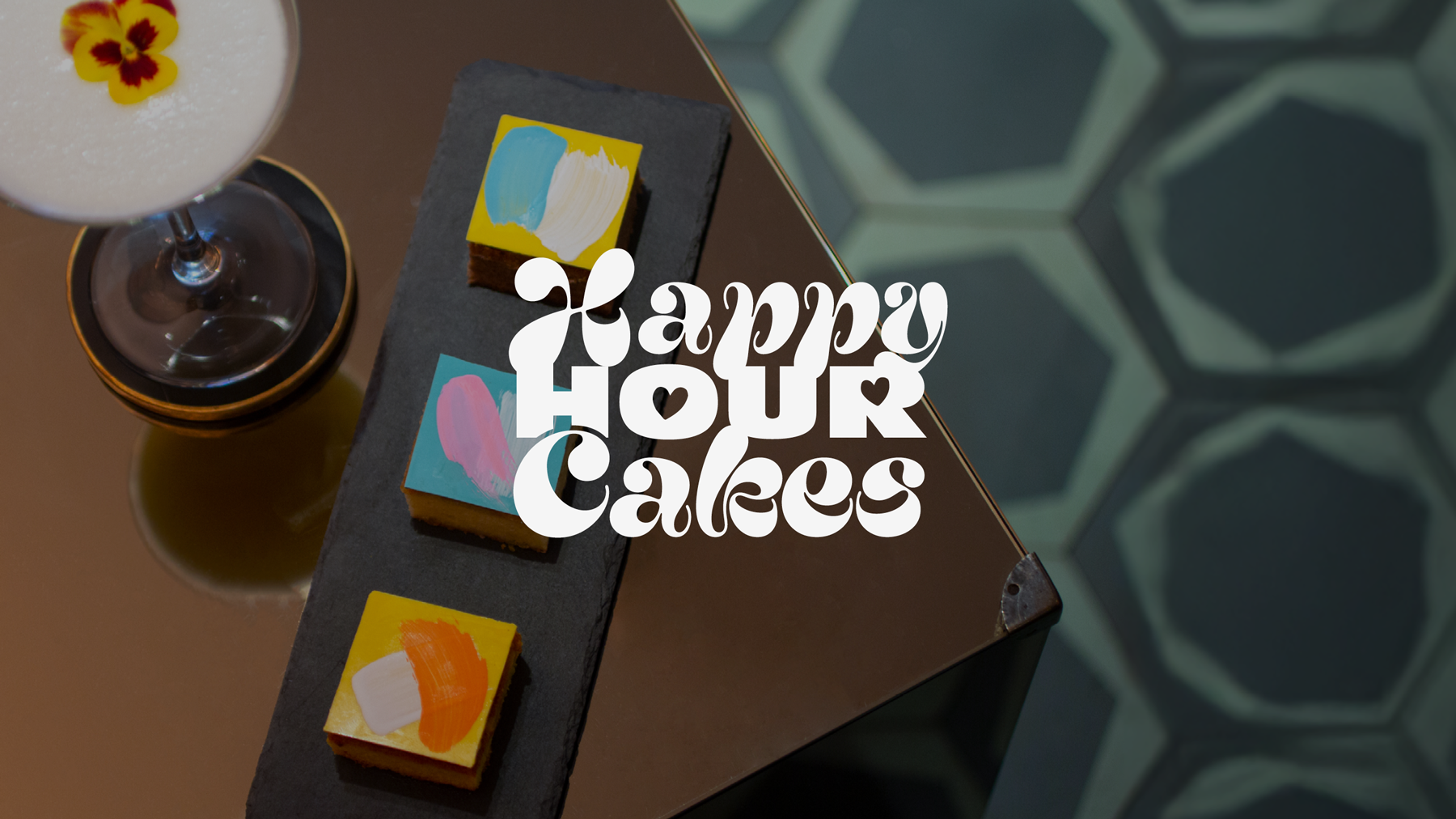 Happy Hour Cakes logo over a moody shot of petit fours and a cocktail.