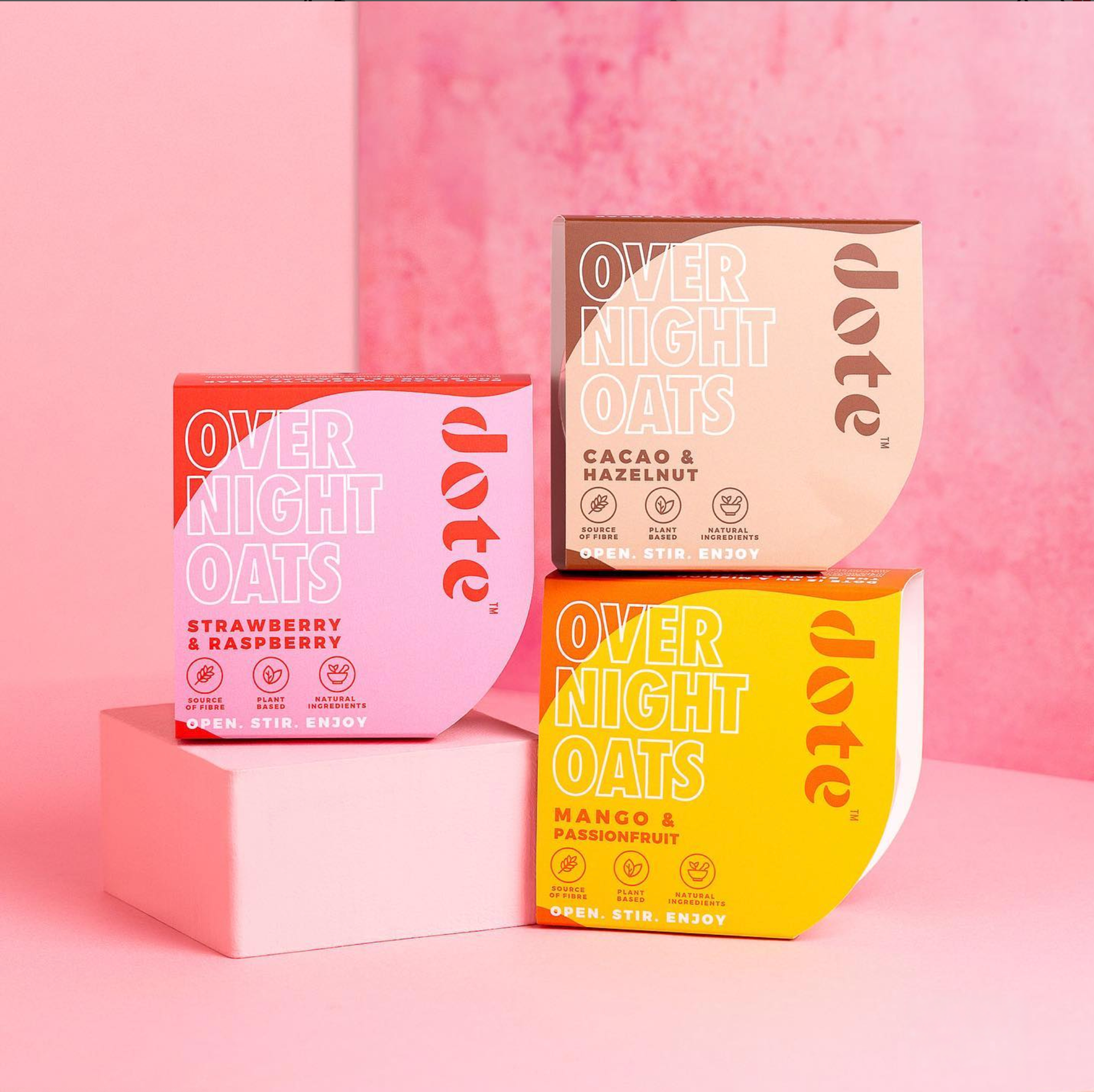 Three Dote overnight oats cartons — strawberry and raspberry, cacao and hazelnut, and mango and passionfruit — stacked on pink plinths against a pink backdrop.
