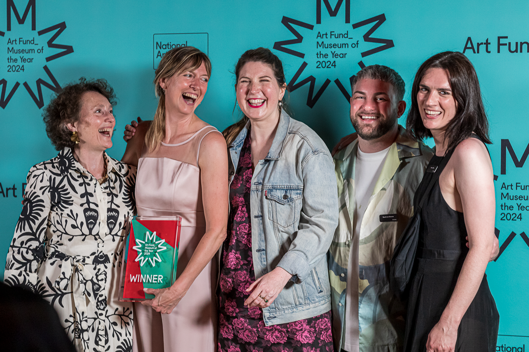 The winning museum team laughing together with the Museum of the Year 2024 trophy in front of a teal branded step-and-repeat.