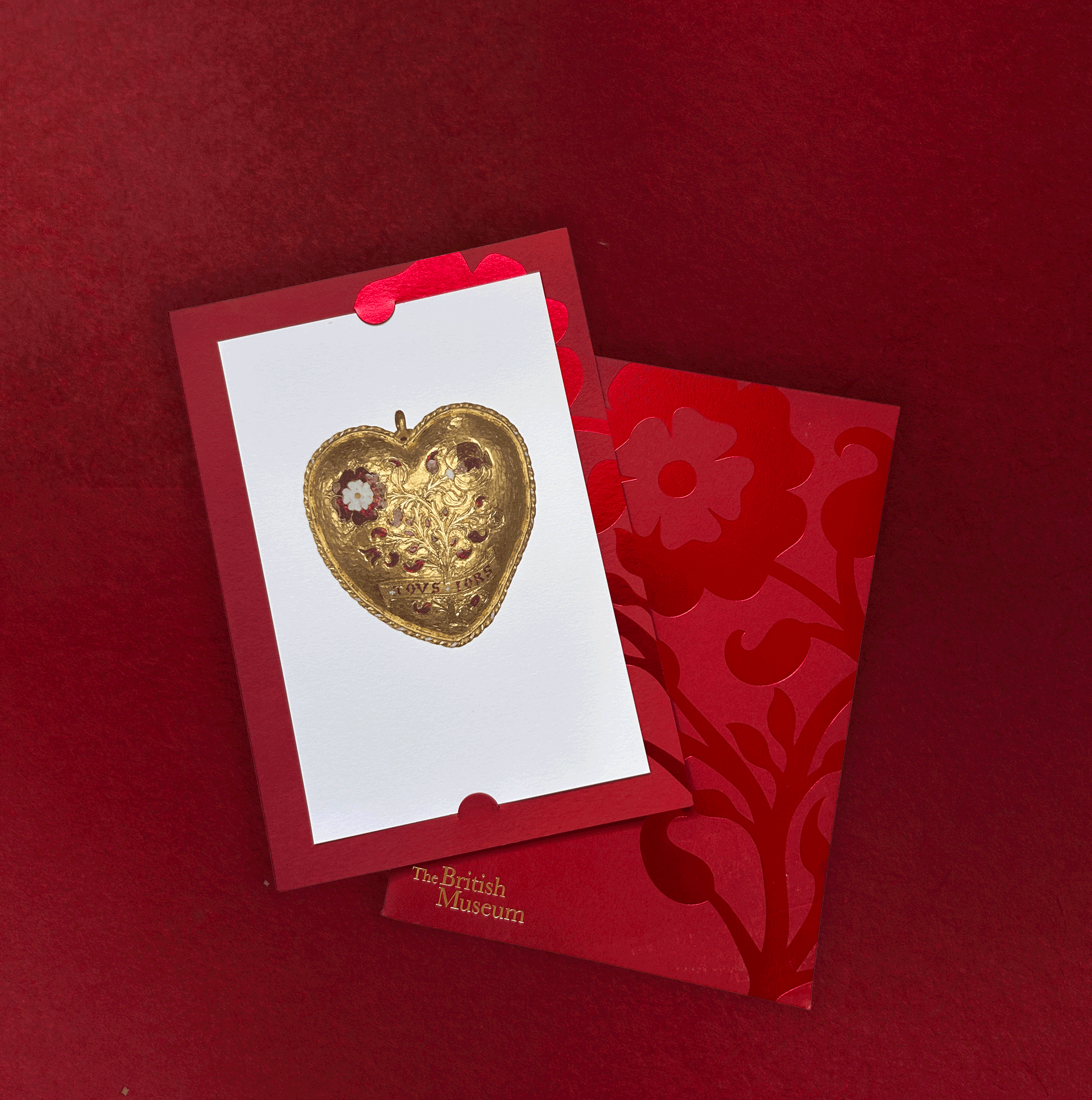 Gold Tudor Heart pendant postcard sitting in its foiled red invitation sleeve on deep red velvet.