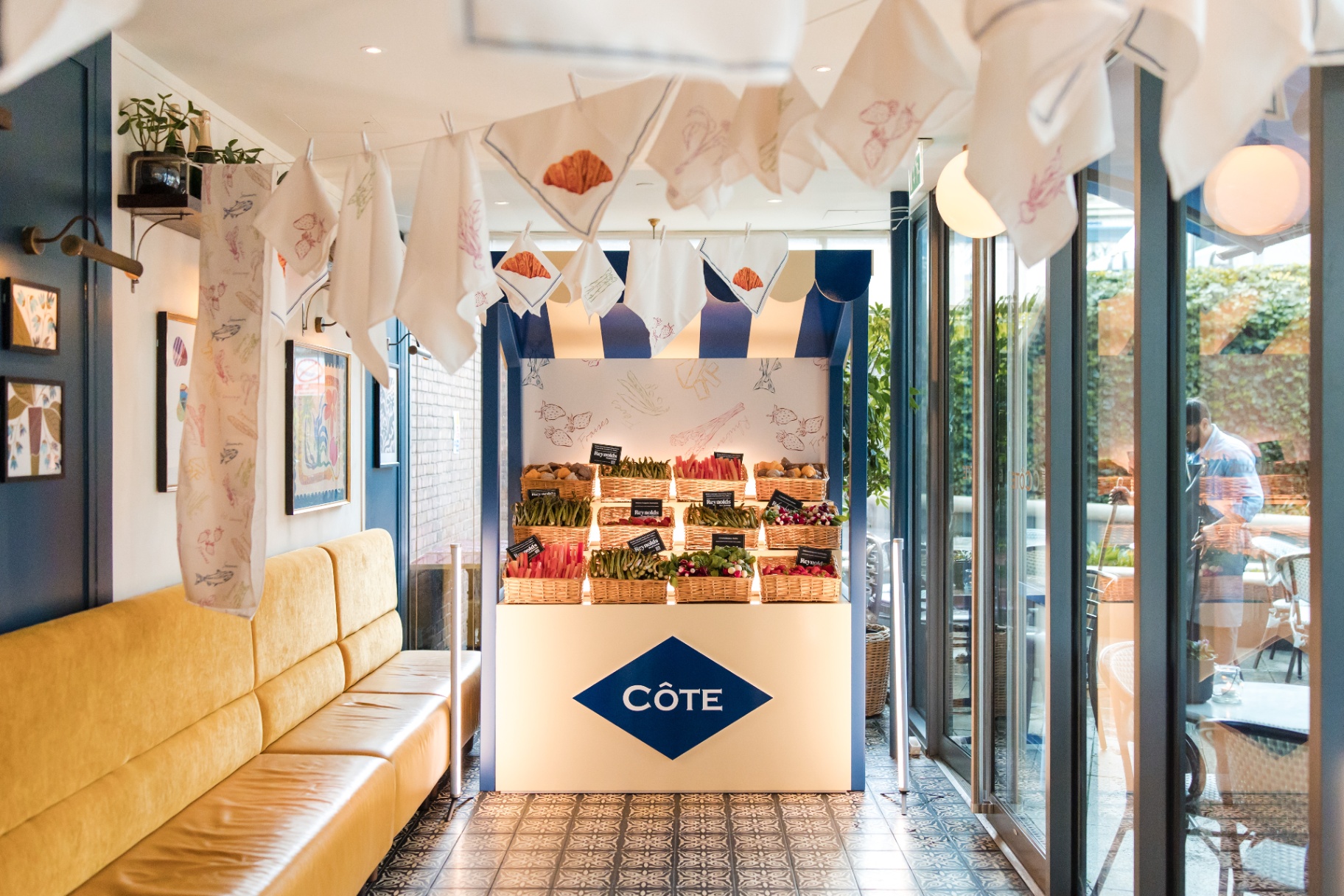 Le Marché market stall installation inside a Côte restaurant, with produce baskets, striped canopy and printed napkins strung overhead.