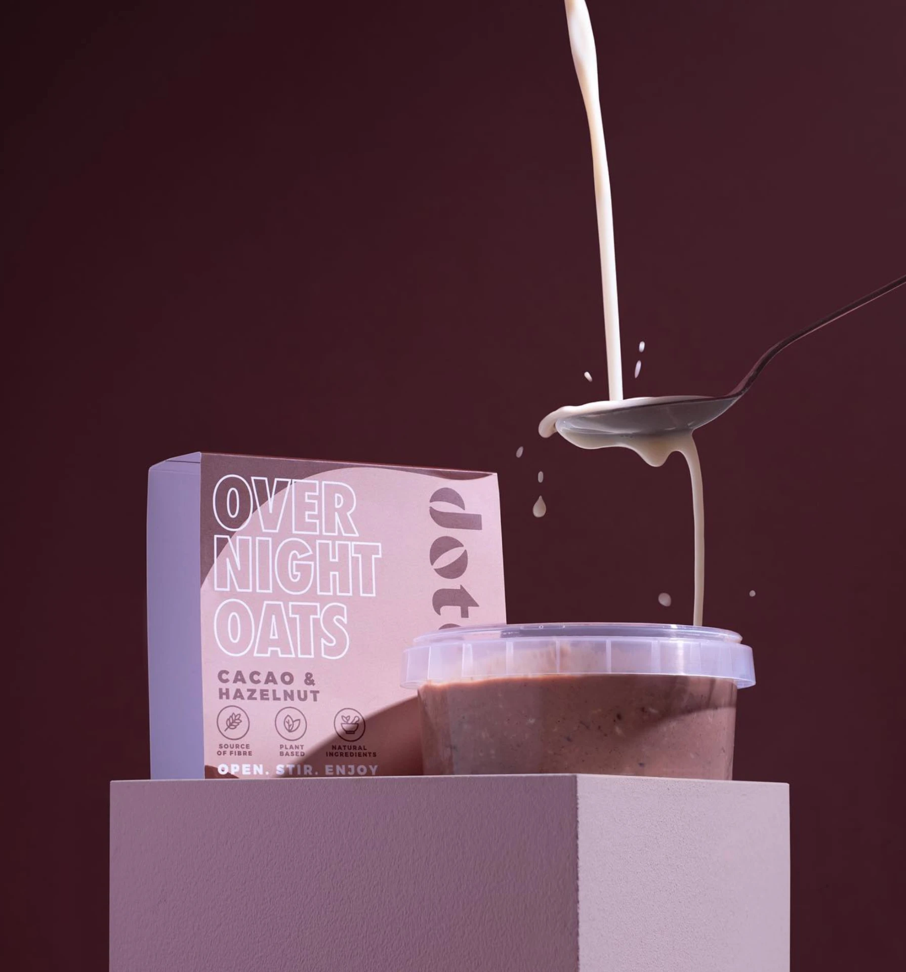 Dote cacao and hazelnut overnight oats pot on a plinth with plant milk pouring over a spoon against a deep burgundy backdrop.